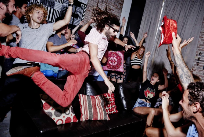 Enthusiastic group of young adults celebrating at an indoor party, one man jumping over a black sofa. Festive atmosphere, casual fashion, indoor setting.