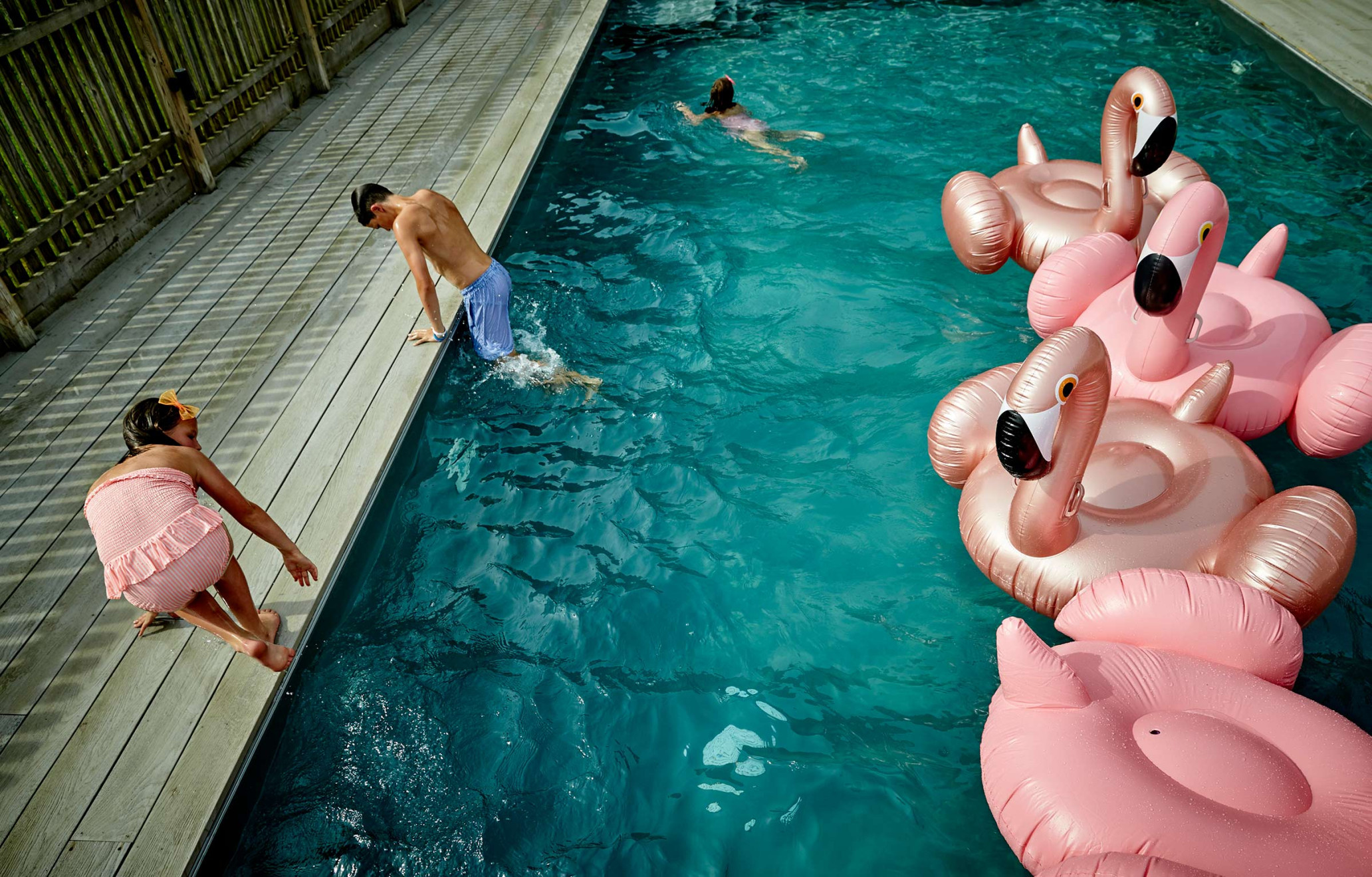 A group of people, including a child, are near a pool filled with water and pink flamingo inflatables, by a wood plank deck and fence.