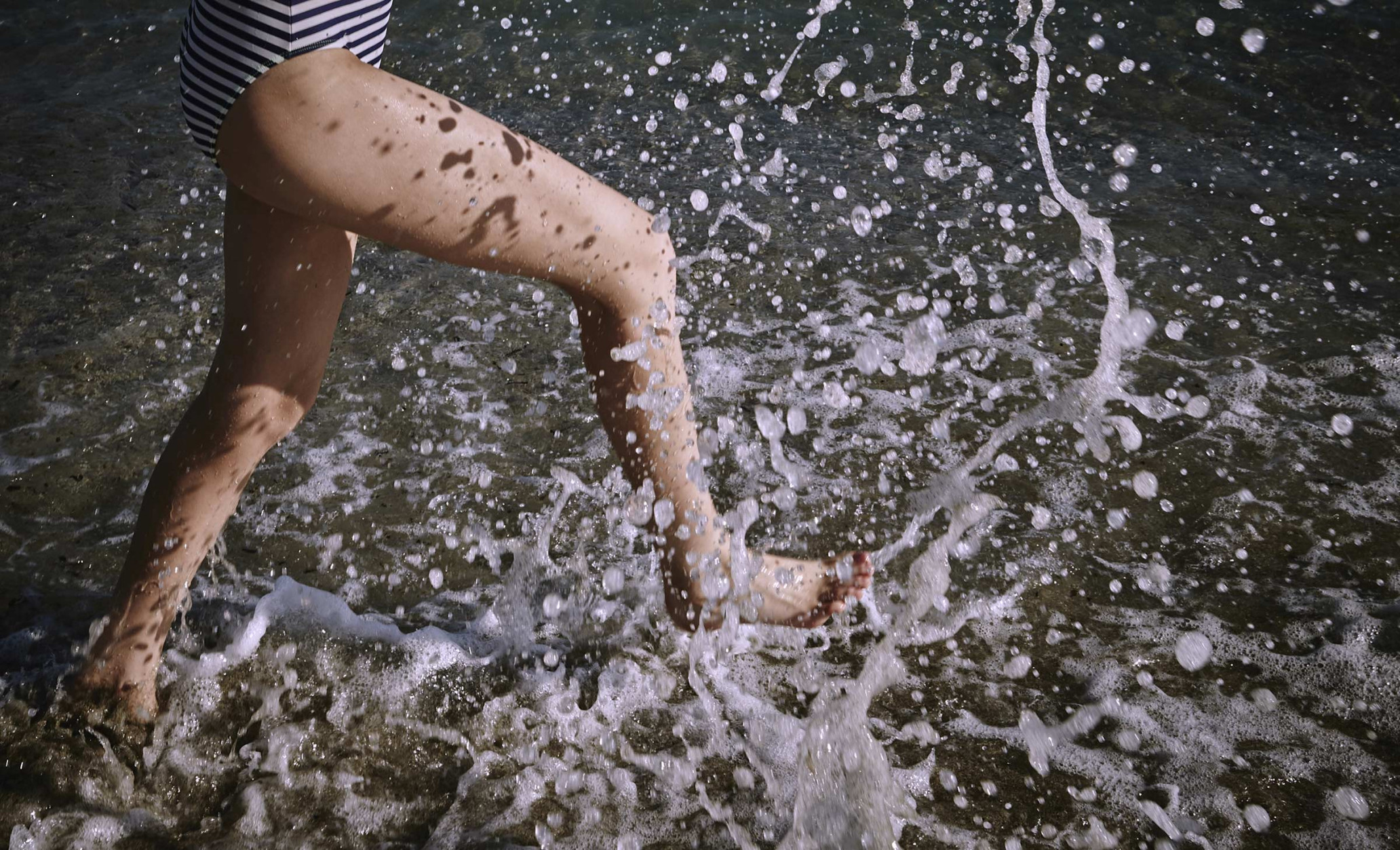 Woman's legs wading in the ocean surf, water splashing. Striped swimsuit visible. Ocean water surrounds the lower legs and splashes upwards.