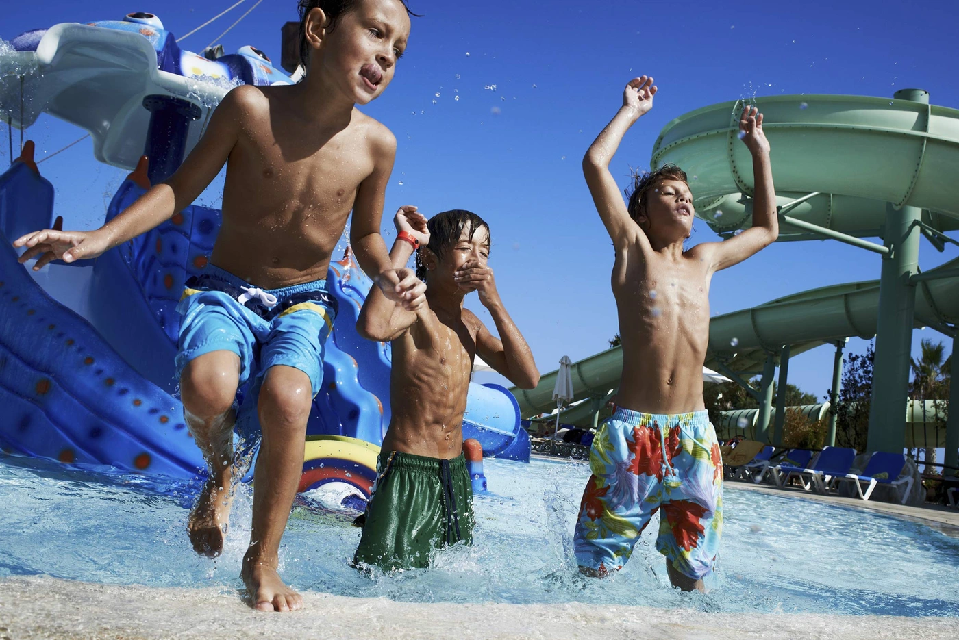 Three boys playing in a sunny outdoor water park with water slides and a pool splashing water.