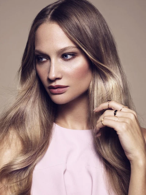Close-up of a blonde woman with blue eyes wearing a pink top and rings on her fingers posing in front of a beige background.