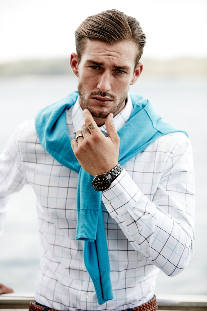 Close-up portrait of a man in his 20s wearing a white plaid shirt and a bright blue sweater draped over his shoulders.