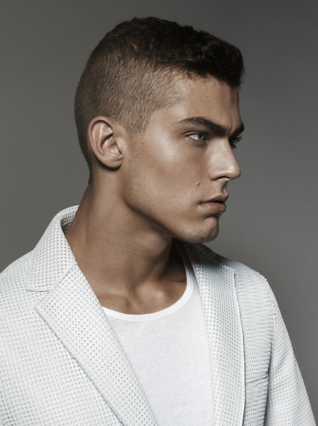 Portrait of a young adult male model with a closely cropped haircut, wearing a white t-shirt and textured blazer jacket against a grey background.