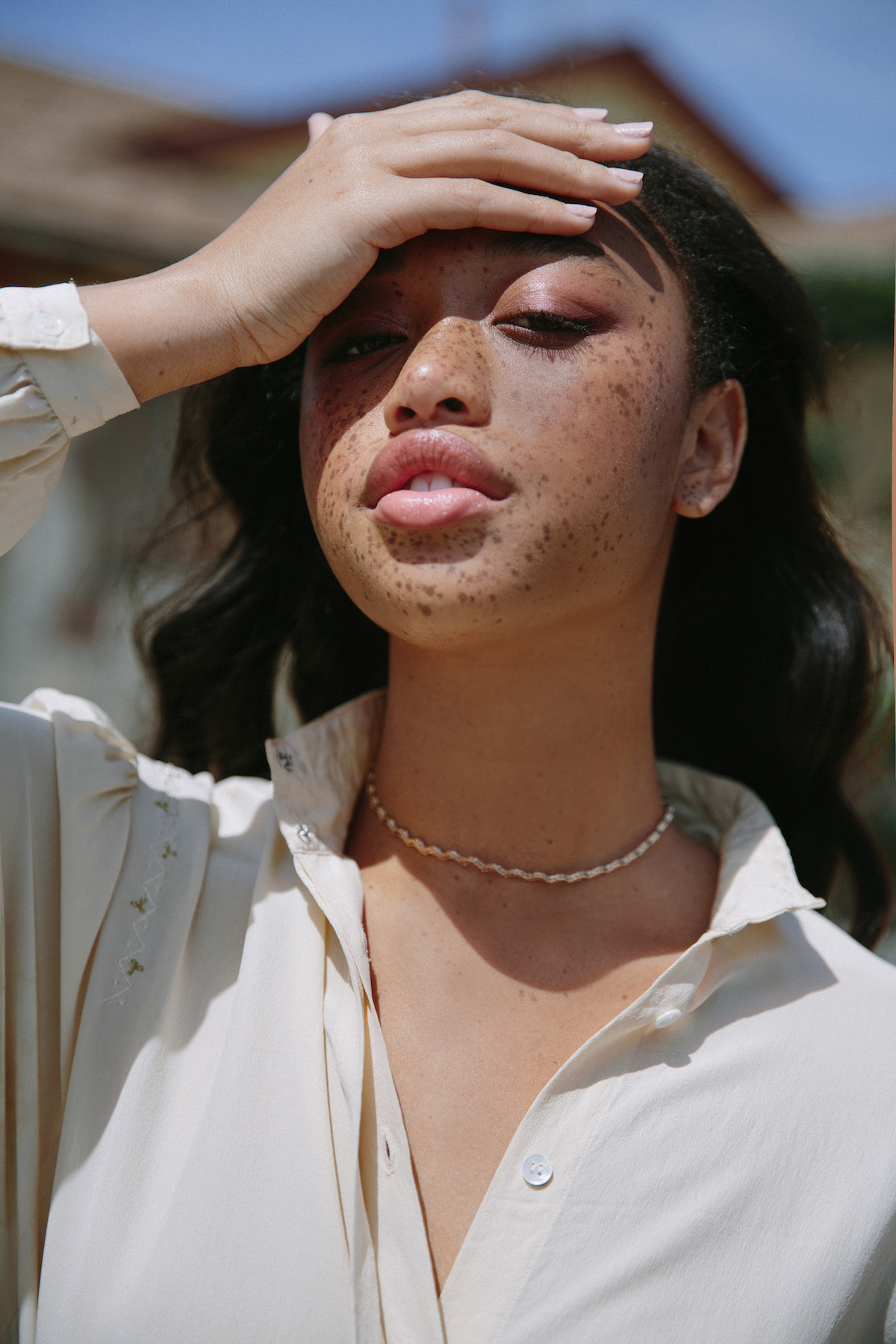 Portrait of a young adult woman with freckles wearing a white button-down shirt and necklace, posing outdoors in bright sunlight.