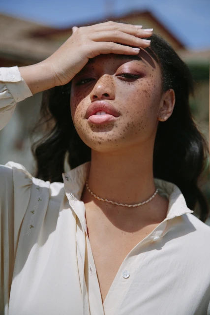 Portrait of a young adult woman with freckles wearing a white button-down shirt and necklace, posing outdoors in bright sunlight.