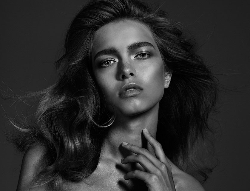 Black and white portrait of a young adult woman with wavy hair, make-up, and hand near her neck, set against a dark gray background.