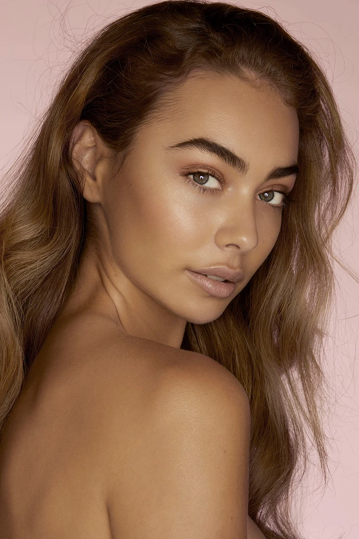 Portrait of a woman with long wavy brown hair and natural makeup looking over her shoulder against a solid pink background.