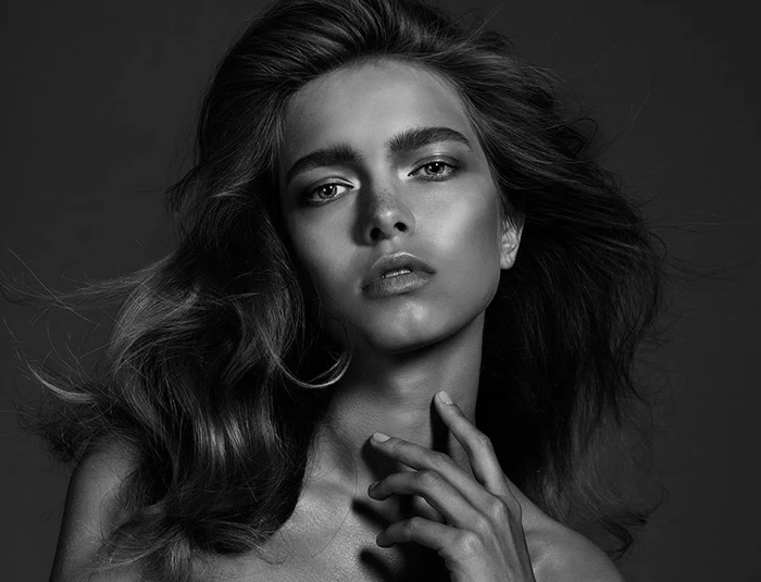 Monochrome portrait of a woman with voluminous wavy hair, touching her neck against a dark background