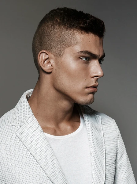 Profile portrait of a man in his 20s with short hair wearing a white textured blazer against a grey backdrop