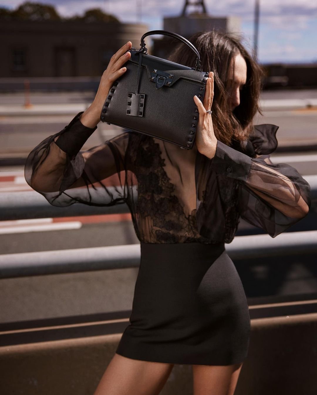 Adult woman wearing a black dress holding a black leather handbag, standing outdoors in an urban setting. Fashion photography.