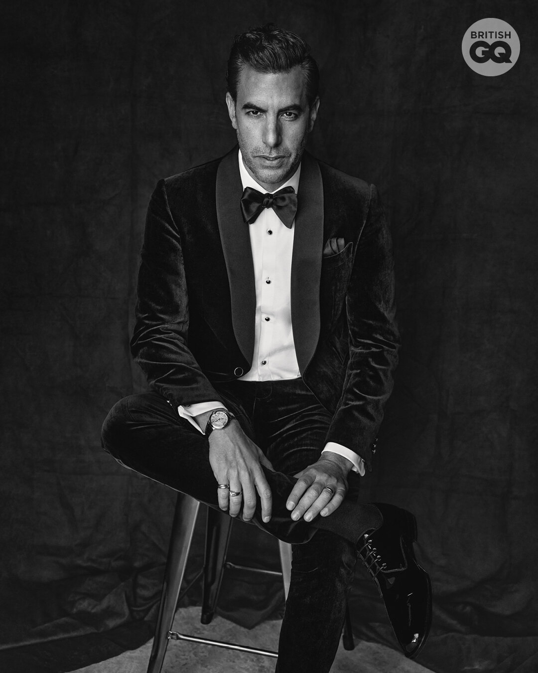 Man in velvet tuxedo sitting on a stool in a black and white studio portrait, wearing a bow tie, wristwatch, and rings.