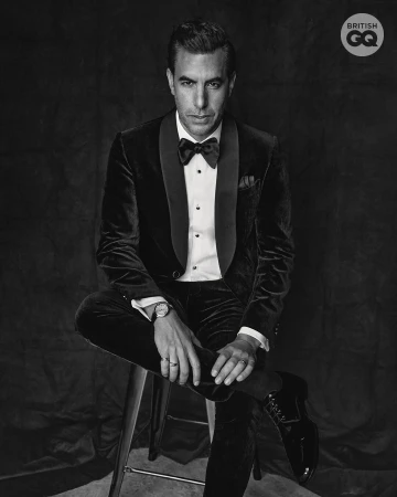 Man in velvet tuxedo sitting on a stool in a black and white studio portrait, wearing a bow tie, wristwatch, and rings.