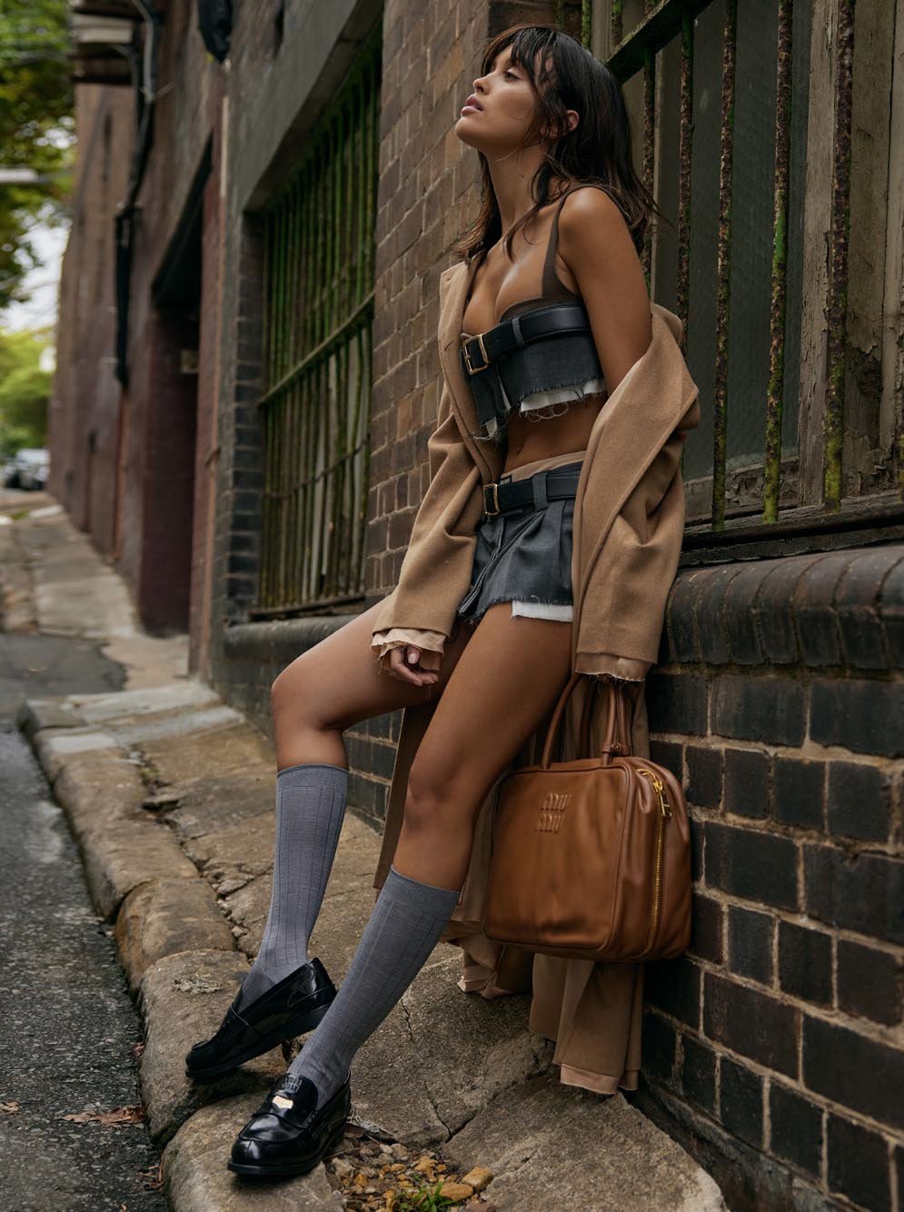 Brunette woman posing outdoors wearing a brown jacket, bustier top, shorts, knee socks, loafers and holding a brown leather purse.