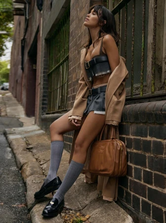 Brunette woman posing outdoors wearing a brown jacket, bustier top, shorts, knee socks, loafers and holding a brown leather purse.