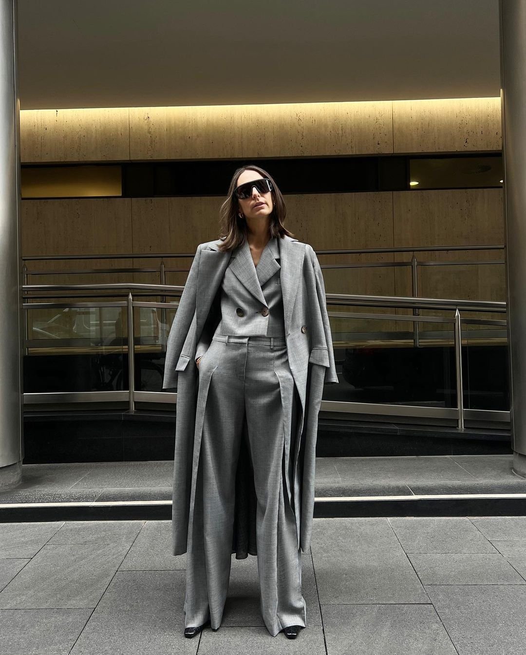 An adult woman wearing sunglasses, a gray suit, and a long gray coat poses indoors with a neutral expression and gray stone background.