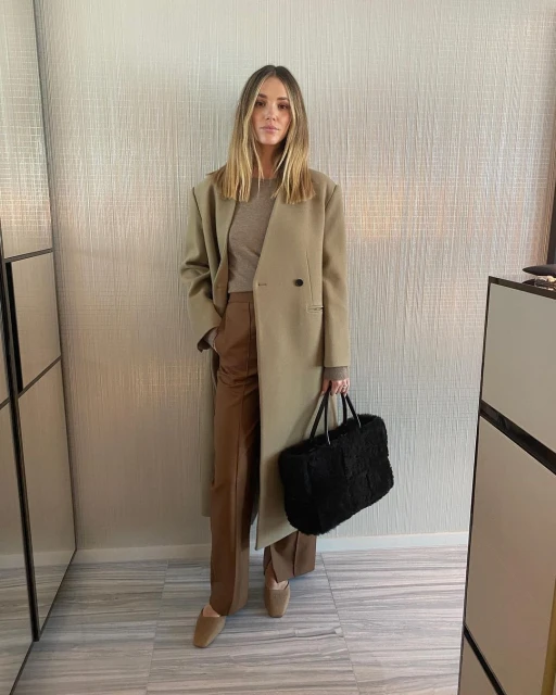 A woman in a tan coat and brown pants stands indoors, holding a black bag, in a fashion-forward pose with neutral tones.