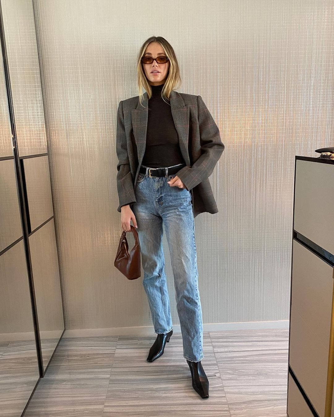 Woman in oversized grey blazer, black turtleneck, and light wash jeans holding a brown leather handbag