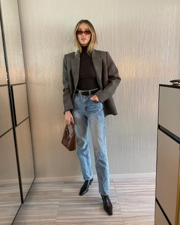 Woman in oversized grey blazer, black turtleneck, and light wash jeans holding a brown leather handbag