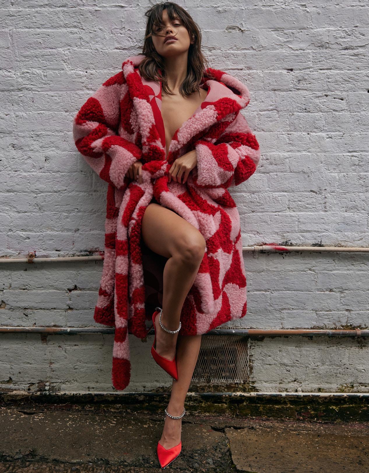 Woman in red and pink patterned coat and red heels poses against a textured brick wall in an outdoor fashion shot.