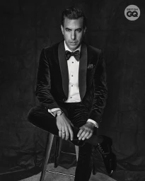 Sacha Baron Cohen seated on a stool wearing a velvet tuxedo and bow tie against a dark textured studio backdrop.