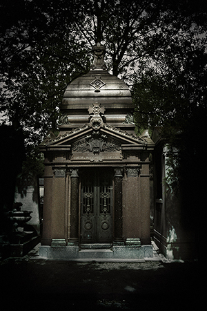 A gothic-style mausoleum stands in a cemetery at night, silhouetted against trees in a dark, moody scene with dramatic lighting.