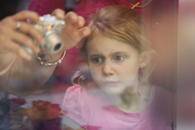 A young girl with blonde hair is the main focus, as someone off-camera takes a photo of her with a small silver digital camera indoors.