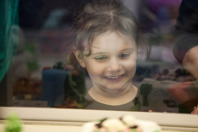 A young child with brown hair smiles and looks down while standing indoors behind a glass partition at an exhibit of some kind.