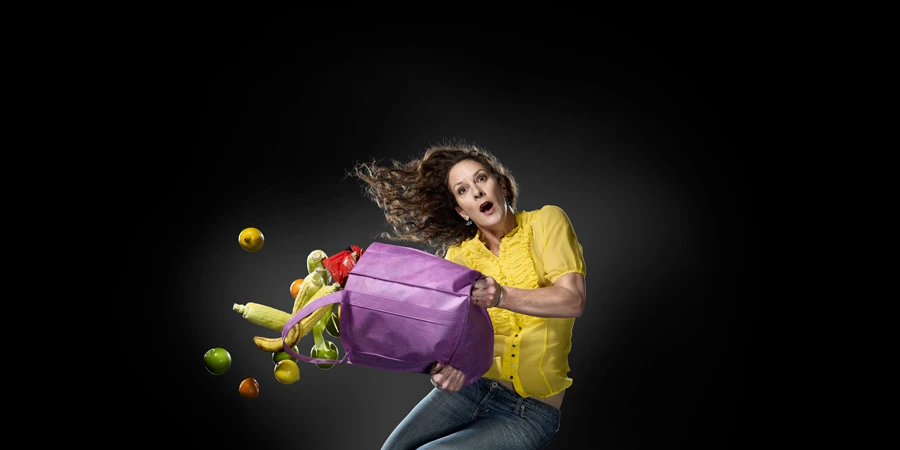 A woman in a yellow blouse and jeans with an open mouth, dropping fruit and vegetables from a purple tote bag against a dark background.
