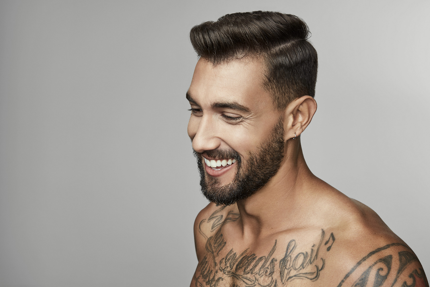 Close-up portrait of a shirtless man with a beard, distinctive haircut, and tattoos smiling, against a neutral grey backdrop.