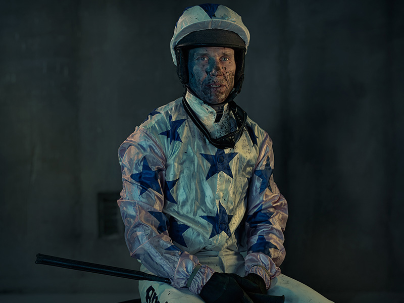 A portrait image features a single jockey in a star-patterned uniform, wearing a helmet and sitting in what appears to be a dark indoor space.