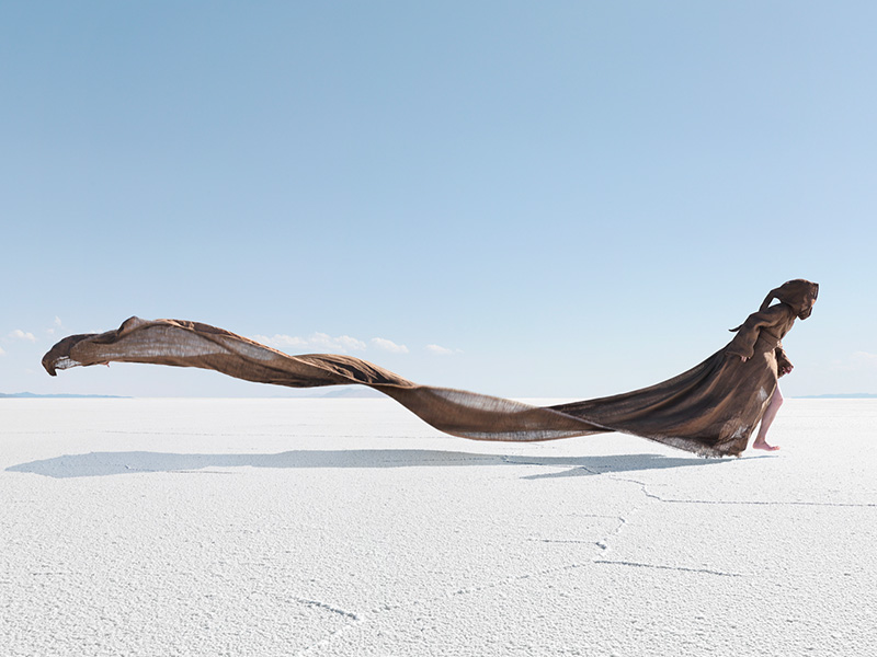 Woman draped in flowing brown fabric walks barefoot across a vast, sunlit salt flat under a clear blue sky in a fashion editorial shot.