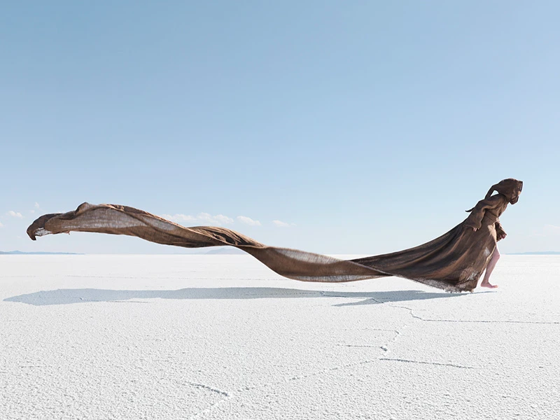 Woman draped in flowing brown fabric walks barefoot across a vast, sunlit salt flat under a clear blue sky in a fashion editorial shot.