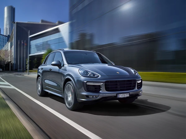 Gray Porsche SUV driving on a city street with motion blur. Modern architecture and buildings are visible in the background.