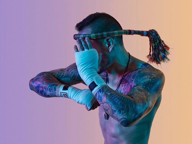Shirtless tattooed man with Muay Thai headwear and wrapped hands posing with a clenched fist and arm raised in a fighting stance.