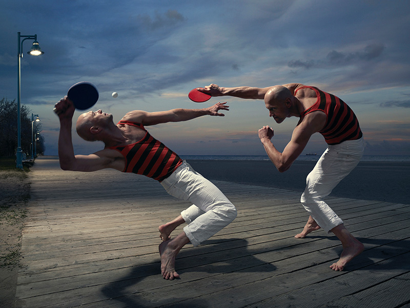 Two bald men playing table tennis barefoot on a wooden boardwalk near the beach against a twilight sky with dramatic clouds.