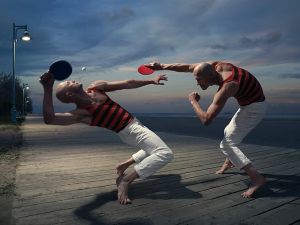 Two bald men playing table tennis barefoot on a wooden boardwalk near the beach against a twilight sky with dramatic clouds.