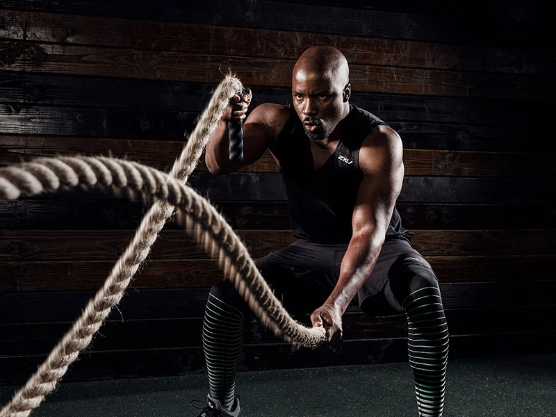 Athletic man exercising with battle ropes in a gym. He is wearing a black tank top and compression tights. The background is a wooden wall.