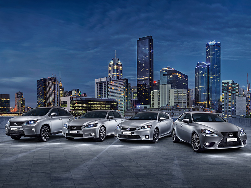Four silver Lexus sedans are parked on a rooftop against a backdrop of city buildings at dusk, showcasing automotive design.