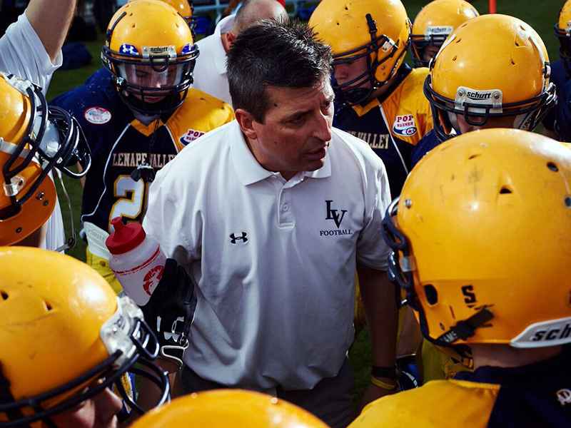 American football coach leading team huddle on field with yellow helmets. Under Armour polo shirt and water bottle, natural light.