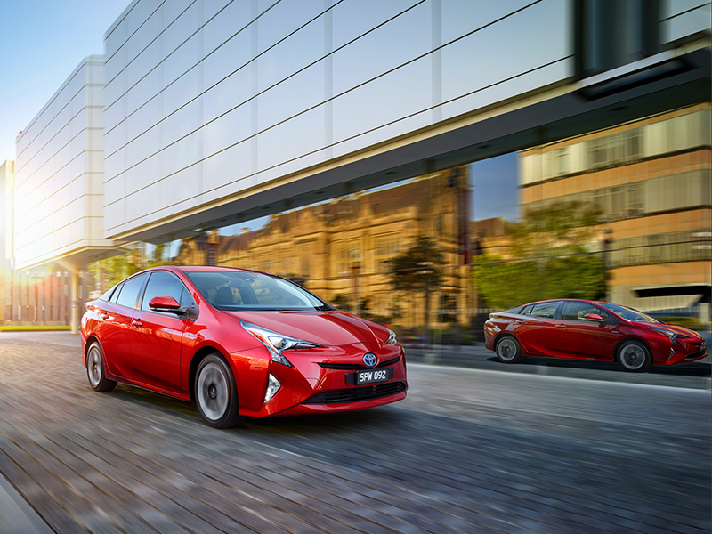 Two red Toyota Prius cars are speeding down a paved road in an urban environment, with modern buildings reflecting the sunlight.