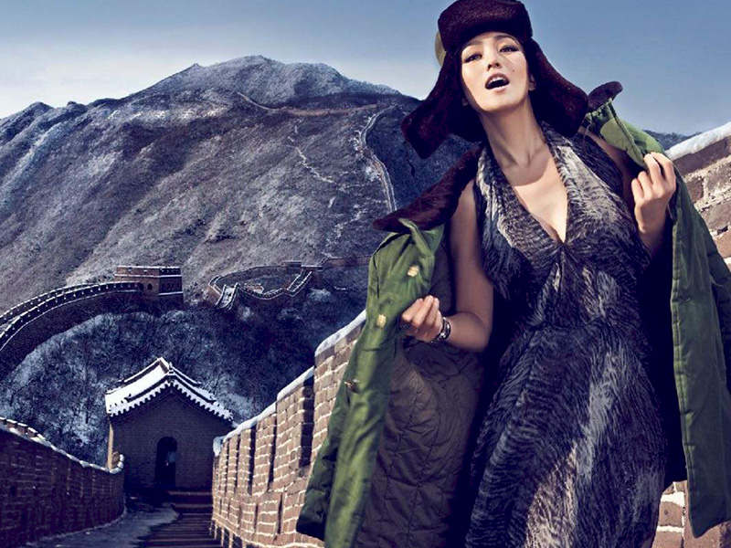 Asian woman wearing hat and patterned dress posing by the Great Wall of China with mountains in background during daylight hours.