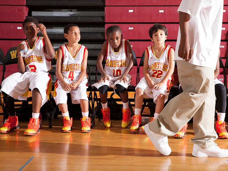 A youth basketball team sits on the bench while the coach walks by in a gym. The kids are wearing white and gold uniforms with orange sneakers.