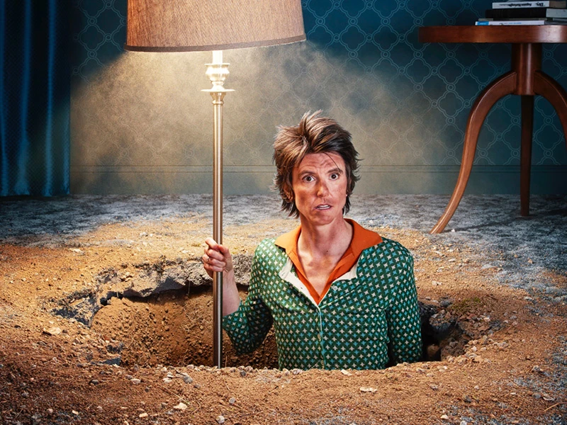 Person in green shirt stands in a dirt hole holding a floor lamp. A table is placed in the background with blue pattern wallpaper.
