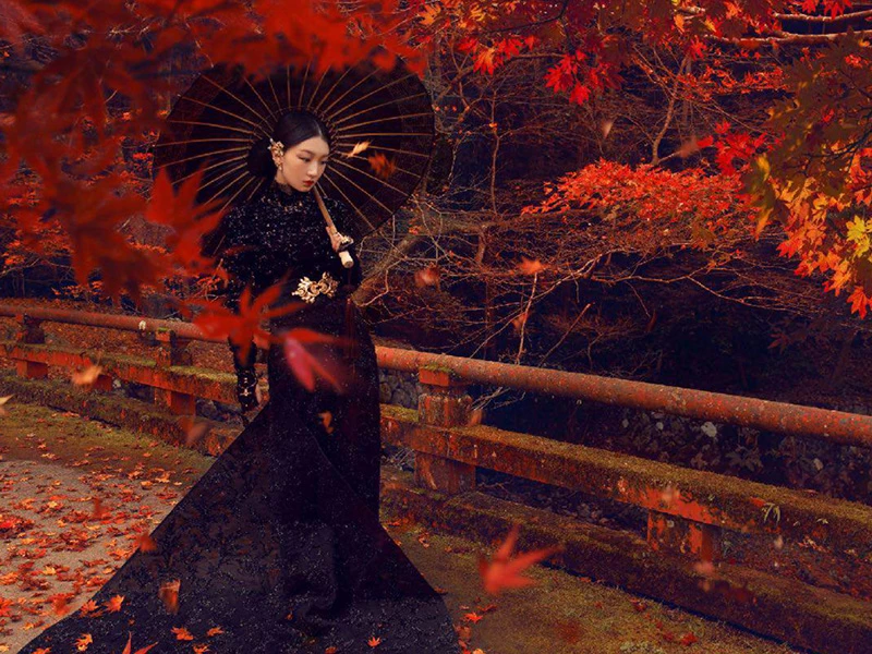 Elegant woman in black gown holding umbrella poses on mossy stone bridge, surrounded by vibrant red and orange autumn leaves.