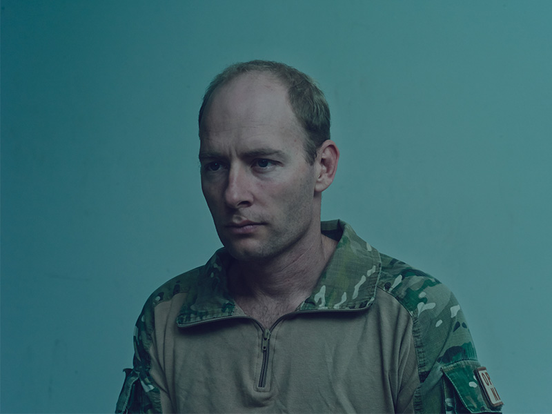 Portrait of an adult male in military camouflage, looking off-camera against a neutral green background in a medium shot.