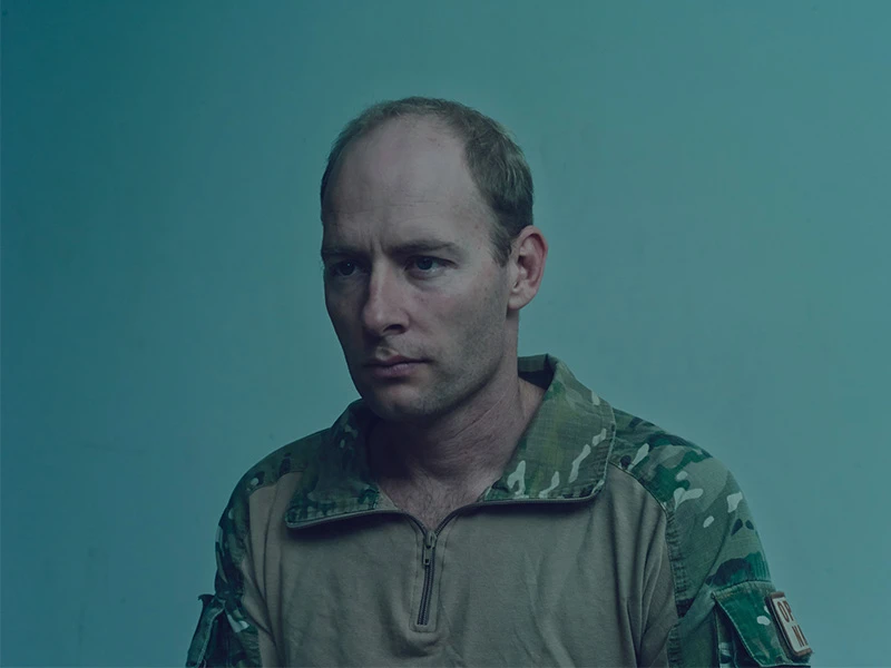 Portrait of an adult male in military camouflage, looking off-camera against a neutral green background in a medium shot.