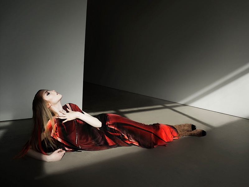 Fashion shot of an adult woman with blonde hair in a flowing red dress lying on the floor in a photo studio with soft directional light.