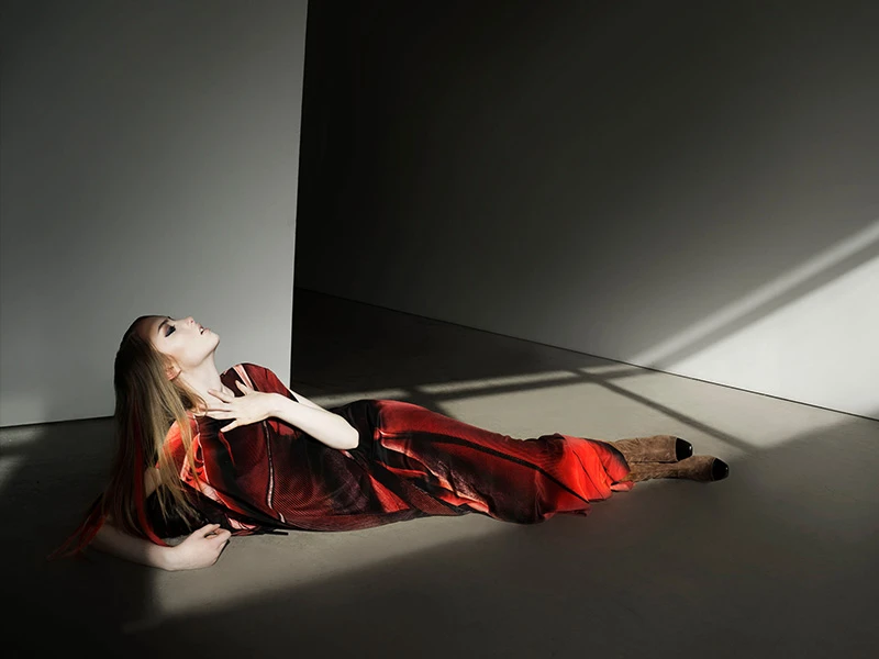 Fashion shot of an adult woman with blonde hair in a flowing red dress lying on the floor in a photo studio with soft directional light.