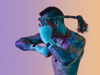 Shirtless male fighter with extensive arm tattoos wearing white hand wraps and a mongkhon headband against a gradient background.