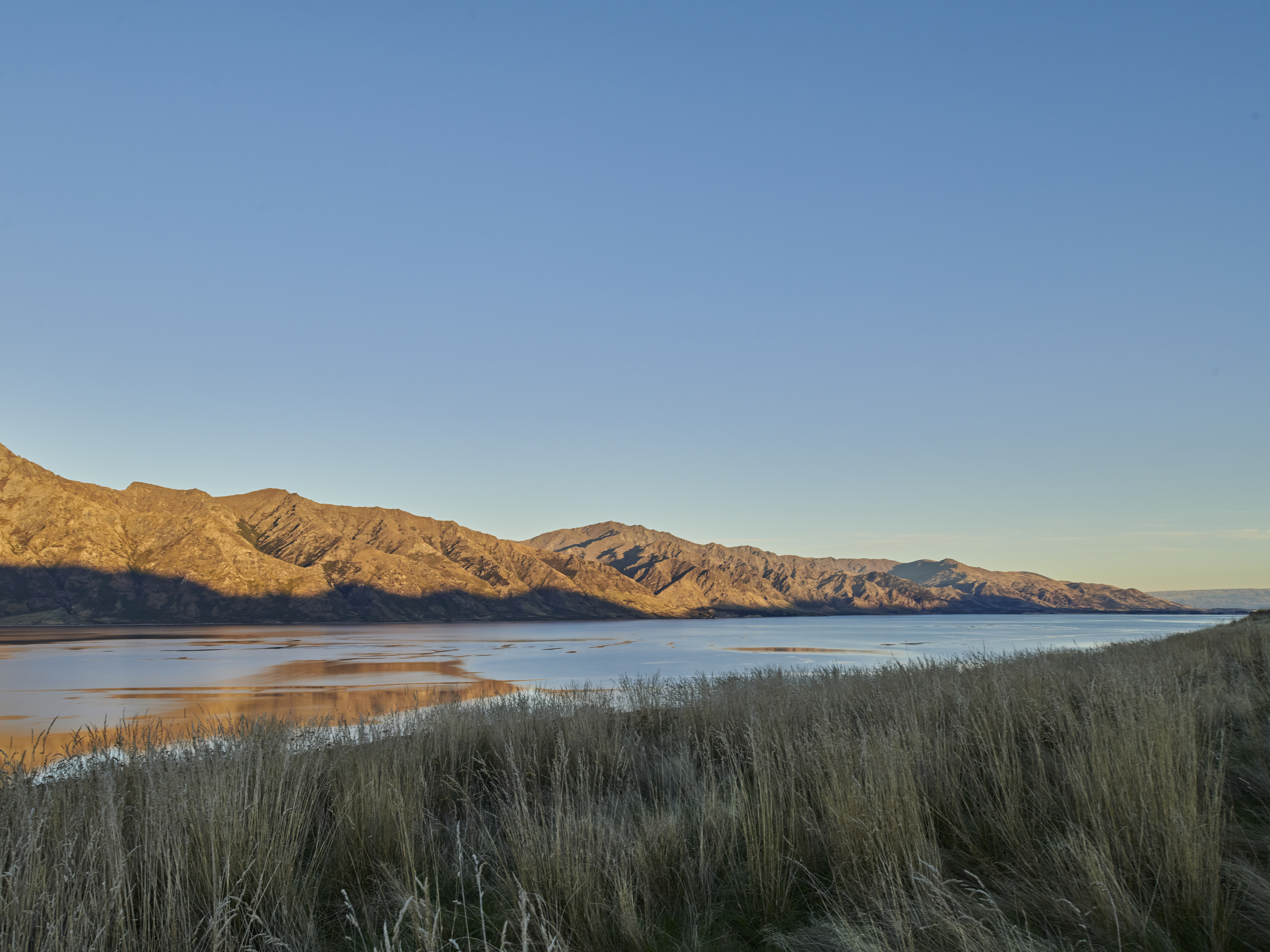 Scenic view of mountains and a lake reflecting the sky at sunset, with golden-brown grasses in the foreground, creating a tranquil landscape.